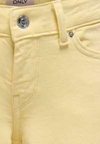 Pantalon en jean jaune clair avec bouton et rivet en métal, poche avant visible, passants de ceinture et étiquette partielle à la taille indiquant "ONLY."