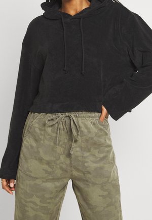 Osoba ubrana w czarną krótka bluzę z kapturem i zielone spodnie moro z troczkami, z jedną ręką w kieszeni, a drugą swobodnie opuszczoną.