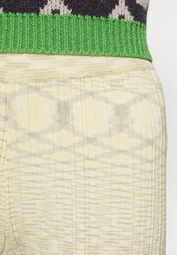 Strikpullover med en grøn ribstrikket halsudskæring, parret med ribstrikkede gule bukser med et subtilt geometrisk mønster i lysere nuancer.