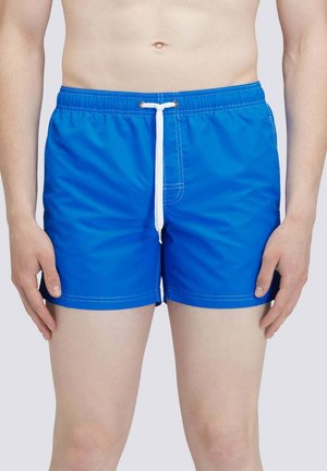 Uomo che indossa pantaloncini da bagno blu brillante con coulisse bianca, in piedi davanti a uno sfondo chiaro e semplice.
