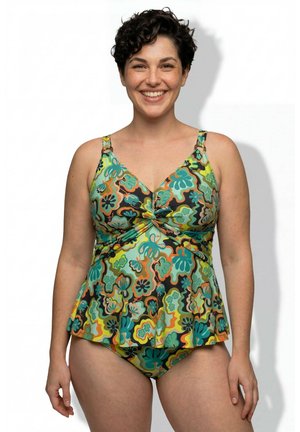 Lächelnde Frau mit kurzen lockigen Haaren, die einen farbenfrohen Tankini mit Blumenmuster vor einem schlichten weißen Hintergrund trägt.