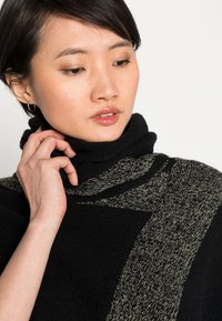 Pull en laine noir à col roulé tricoté avec des accents dorés texturés, présentant un haut col et un design diagonal unique sur le devant.