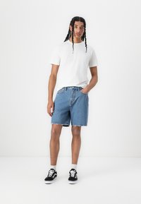 Tricou alb din bumbac, pantaloni scurți din denim albastru până la genunchi, cu tiv desfăcut, adidași negri cu talpă albă, poză în picioare pe un fundal simplu.