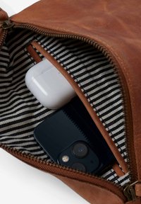 Braune Lederhandtasche mit schwarzem Smartphone und weißem Etui für drahtlose Ohrhörer im Inneren, mit gestreifter Stoffauskleidung und teilweise geöffnetem Reißverschluss.
