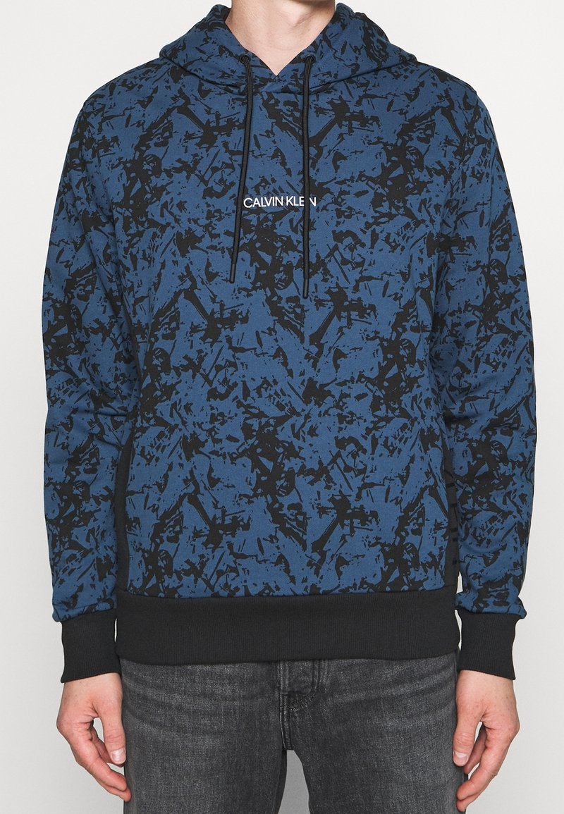 Sweat à capuche à motif abstrait bleu et noir avec poignets et ourlet noirs, logo « Calvin Klein » centré sur la poitrine, porté avec un jean gris foncé.