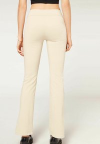Pantaloni beige a zampa d'elefante con una texture liscia, vita alta e dettagli di cucitura minimi, abbinati a scarpe nere.