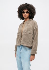 Žena s kučeravými vlasmi nosiaca béžovú fleecovú bundu, slnečné okuliare, hnedejšiu košeľu a svetlomodré džínsy na jednoduchom bielom pozadí.