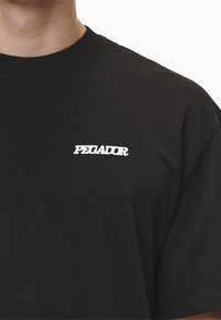 Camiseta de algodón negra con mangas cortas, que presenta un logo bordado en blanco que dice "PEGADOR" en el pecho, textura suave y corte clásico.