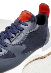 Navy blue sneaker with leather and suede panels, dark blue laces, white sole, and bright orange suede tongue and heel tab.