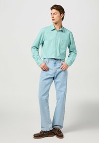 Camisa de cuadros verde de botones, jeans de denim azul claro, zapatos de cuero marrón. Ajuste slim con mangas largas y un detalle de bolsillo en el pecho.