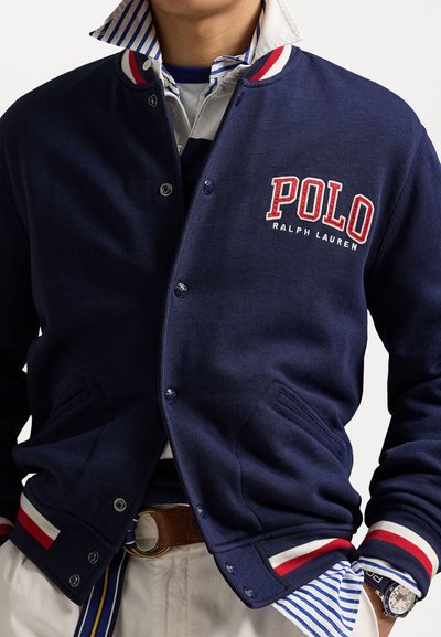 Jaqueta varsity azul marinho com punhos canelados listrados em vermelho e branco, logo da POLO, botões de pressão e bolsos laterais, sobre uma camisa listrada.