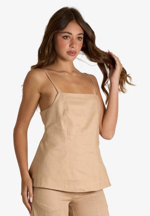 Giovane donna con lunghi capelli castani indossa un top beige senza maniche e pantaloni abbinati, tenendo una ciocca di capelli con una mano.