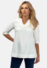 Biała bluzka z 3/4 rękawem, z dekoltem w kształcie litery V, wykonana z lekkiego materiału z bocznymi rozcięciami. Posiada subtelny blask i akcent w postaci małego logo.
