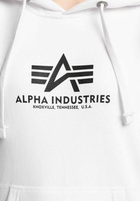 Biela mikina s kapucňou a čiernym potlačeným logom so štýlovými písmenami a textom "ALPHA INDUSTRIES" pod ním. Šnúrky na kapucni.