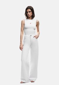 Vrouw in een witte mouwloze top met een rode chili-print en hoge, wijde witte broek, staand met één hand in de zak.