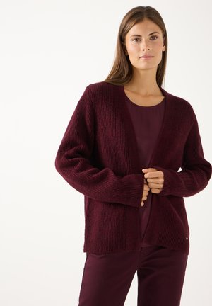 Vrouw draagt een donker bordeauxrode gebreide cardigan over een bijpassende top en broek, staande tegen een effen lichte achtergrond.