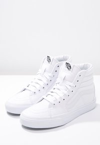 White high-top sneakers made of canvas with reinforced toe, lace-up design, padded collar, and distinctive logo on the tongue.