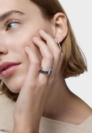 Donna con capelli corti che si tocca la guancia, indossa anelli d'argento con pietre nere alle dita e un orecchino a cerchio d'argento.