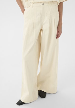 Krémenové nohavice s širokými nohavicami, prednými záhybmi a rozparkami, v páre s čiernymi topánkami so štvorcovou špičkou, nosené osobou s rukami v vreckách.