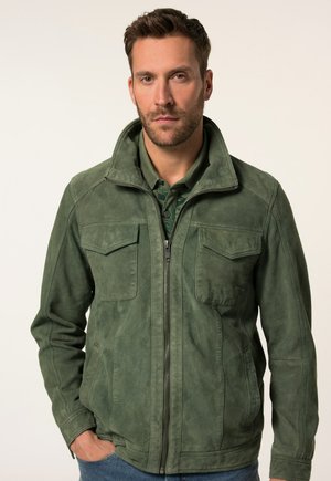Chaqueta de ante verde con cierre de cremallera, que presenta dos bolsillos en el pecho, un cuello alto y un acabado texturizado. Lleva puesta sobre una camisa oscura.