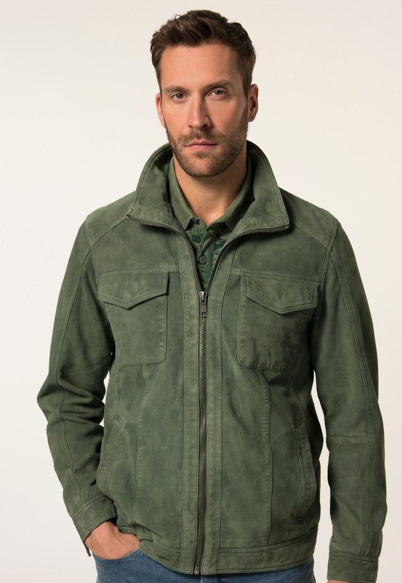 Grüne Wildlederjacke mit einem Reißverschluss, zwei Brusttaschen, einem hohen Kragen und einer strukturierten Oberfläche. Getragen über einem dunklen Hemd.