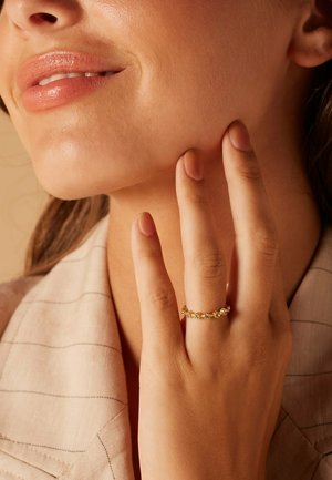 Gold ring with multiple small gemstones, featuring a smooth, shiny surface. Hand gently touching face, wearing a light-colored fabric.