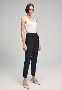 Zwarte op maat gemaakte broek met een hoge taille, voorplooien en een gouden knoop. Gecombineerd met een fitted witte geribde tanktop en lichtgrijze hakschoenen.