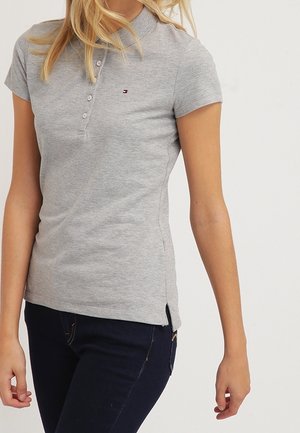 Vrouw draagt een nauwsluitend lichtgrijs polo shirt met korte mouwen en knopen, gecombineerd met donkerblauwe jeans, staand tegen een effen achtergrond.