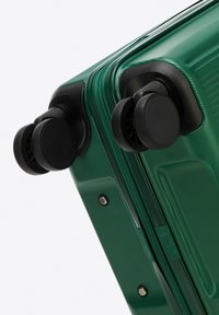 Green hard-shell suitcase with a textured finish, featuring black swivel wheels and a zipper closure. Accents include metal studs and seams.