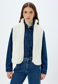 Weißer Kunstpelz-Westen mit hohem Kragen, Reißverschluss und abgerundeten Kanten, kombiniert mit einem blauen Denim-Hemd und hochgeschnittenen Jeans.
