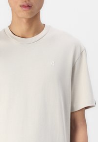 Lys beige t-shirt lavet af bomuld, med rund halsudskæring, korte ærmer og et lille broderet logo på venstre bryst.