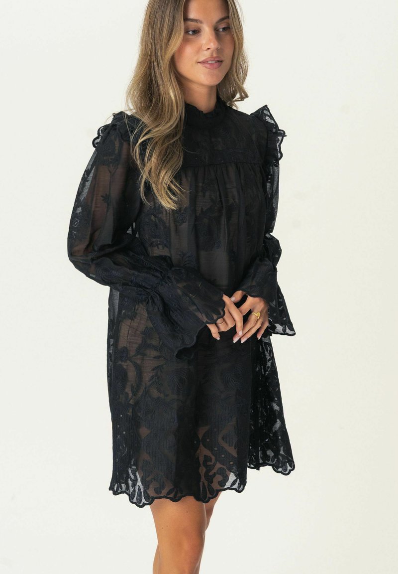 Schwarzes Spitzenkleid aus durchsichtigem Stoff, mit Rüschensöcken und floralem Muster. Der hohe Kragen und der gewellte Saum setzen Akzente.
