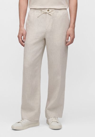 Personne portant un pantalon en lin beige avec taille élastique et cordon, associé à des baskets écru sur un fond uni.
