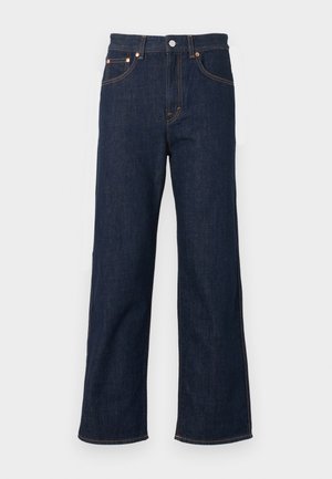 Person iført en lys orange T-shirt med korte ærmer, mørkeblå jeans med lige ben, sorte sko, der holder en sort håndtaske mod en ensfarvet baggrund.