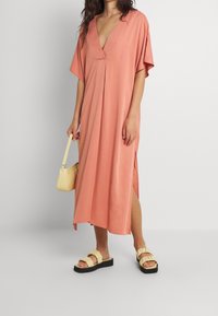 Pfirsichfarbenes Midi-Kleid mit tiefem V-Ausschnitt, kurzen Ärmeln und seitlichen Schlitzen. Getragen mit einer gelben Handtasche und klobigen, rüschierten Sandalen.