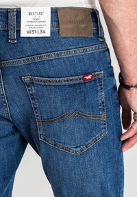 Ciemnoniebieskie dżinsy z denimu o luźnych nogawkach i niskim stanie. Posiadają pomarańczowe przeszycia, brązową skórzaną łatkę oraz małe czerwone logo na tylnej kieszeni.