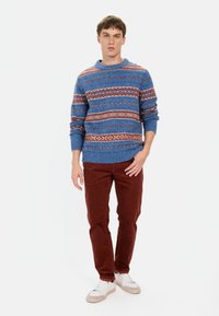 Blauer Strickpullover mit mehrfarbigen Streifen und Mustern. Kombiniert mit kastanienbraunen Hosen und weißen Sneakers. Lässiger Schnitt.