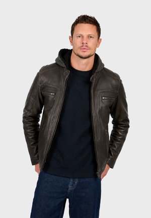 Homme portant une veste en cuir marron foncé avec des poches zippées sur un pull noir et un jean bleu, debout les mains dans les poches.