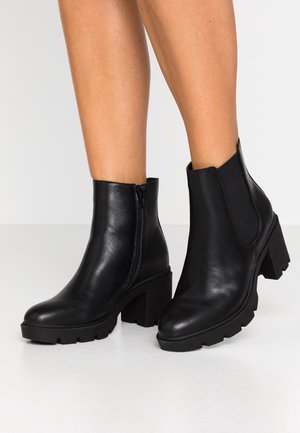 Bottes de cheville en cuir noir avec talons carrés, fermeture éclair latérale sur la botte gauche, panneau élastique sur la droite, portées sur jambes nues sur un fond uni.