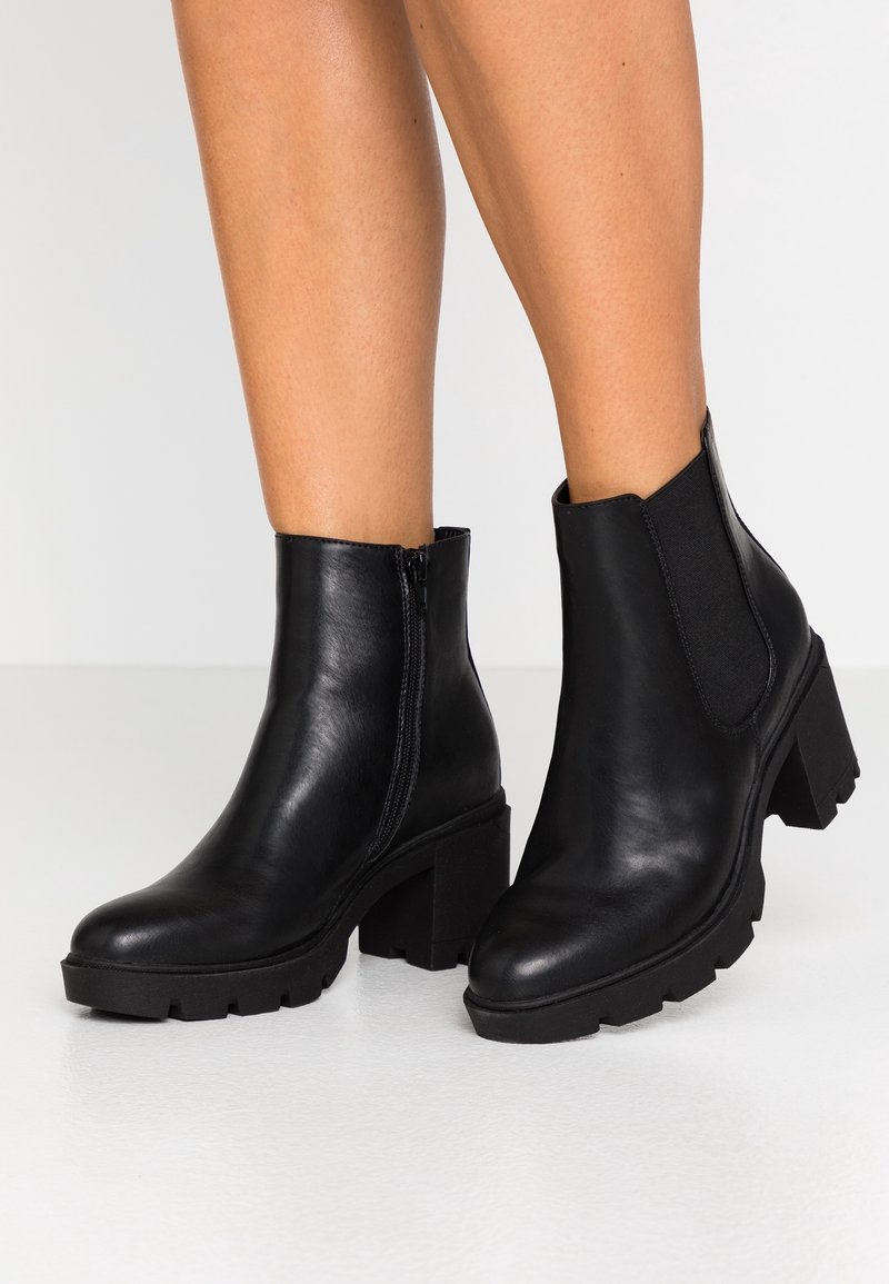 Bottes de cheville en cuir noir avec talons carrés, fermeture éclair latérale sur la botte gauche, panneau élastique sur la droite, portées sur jambes nues sur un fond uni.