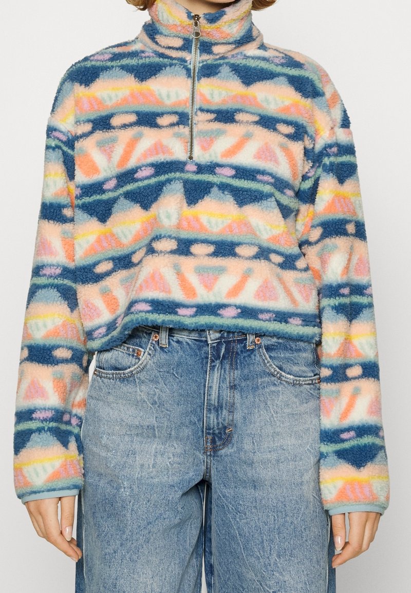 Pull duveteux multicolore à motifs avec demi-fermeture éclair, associé à un jean bleu taille haute, coupe ample, porté par une personne debout.