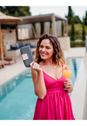 Schwarze Brieftasche mit pinkfarbenem Verschlussdetail, die in der Hand gehalten wird. Im Hintergrund sind ein Schwimmbecken und eine stilvolle Außenlounge zu sehen.
