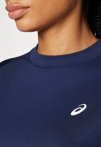 Nahaufnahme einer Person, die ein dunkelblaues Oberteil mit weißem ASICS-Logo auf der Brust trägt und ihre Lippen, Kinnlinie und den Hals zeigt.