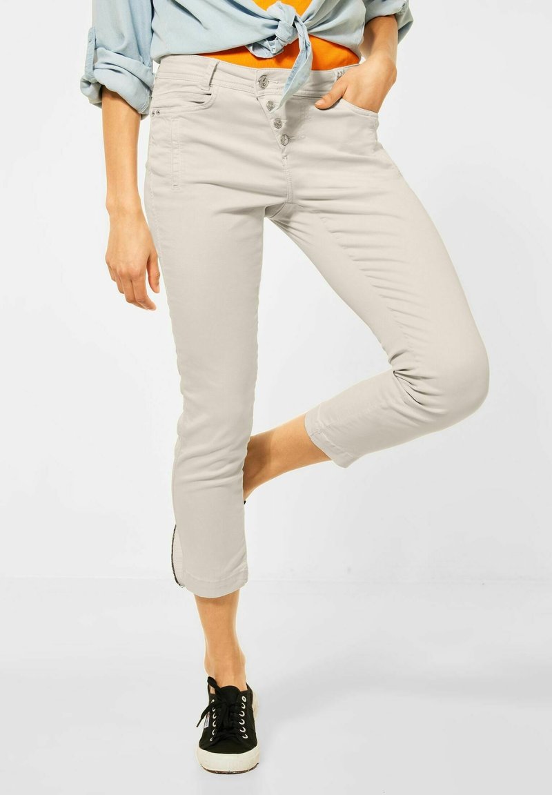 Personne portant un pantalon beige clair coupé court, une chemise orange, un chemisier bleu clair noué et des baskets noires, avec une main dans une poche.