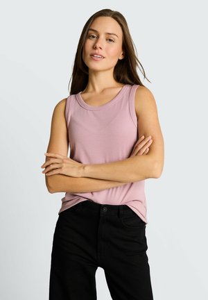 Młoda kobieta o długich brązowych włosach, ubrana w różowy top bez rękawów i czarne spodnie, stojąca z założonymi rękami na jednolitym tle.