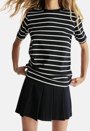 Femme portant un t-shirt manches courtes rayé noir et blanc et une jupe plissée noire, ajustant l'ourlet de son t-shirt.
