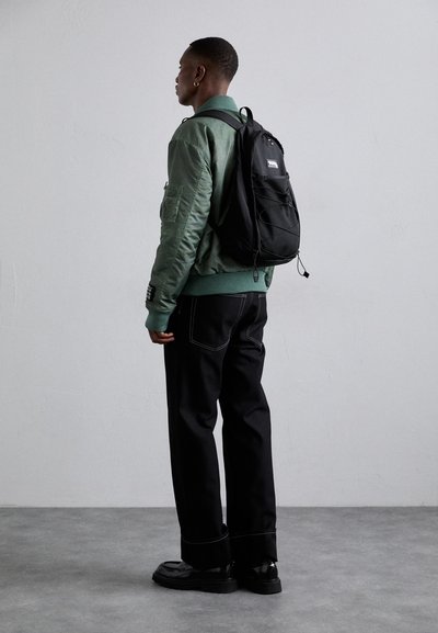 Veste bomber verte avec des poignets côtelés, sac à dos en maille noire, pantalon large noir avec des coutures blanches, et chaussures noires à semelles épaisses.
