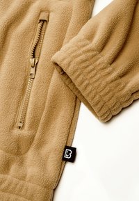 Manche et poche avant zippée d'une veste en polaire beige, avec poignets côtelés et petite étiquette de marque noire en tissu sur fond blanc.