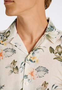 Camisa de botones de manga corta en tejido ligero, con un fondo color crema y patrones florales en tonos pastel y acentos de hojas verdes.