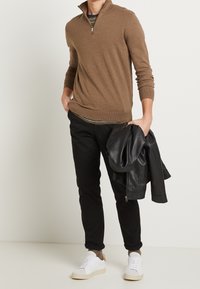 Man som bär en brun tröja med kort dragkedja, svarta byxor och vita sneakers, håller en svart läderjacka i ena handen.